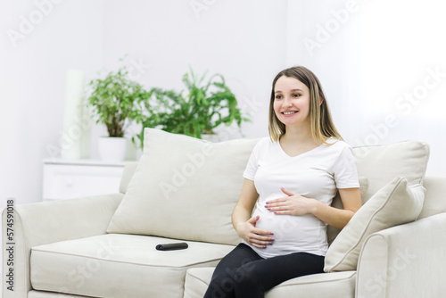 Pregnant woman sitting on white sofa and touching her stomach.