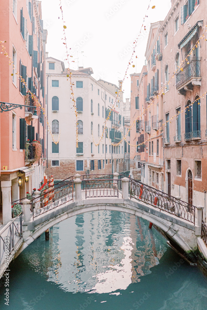 A canal runs through the canyons of houses in Venice, a beautiful bridge connects the sides. The water glitters turquoise and the sun is reflected in the brick facades of the houses.