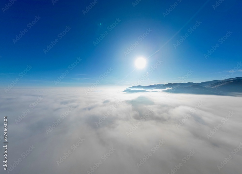 Panoramic aerial drone wide angle view above the sea of clouds with a deep blue sky, a beautiful radiant sun and some mountains and hills, a fairy tale-like dreamy cotton bed at altitude