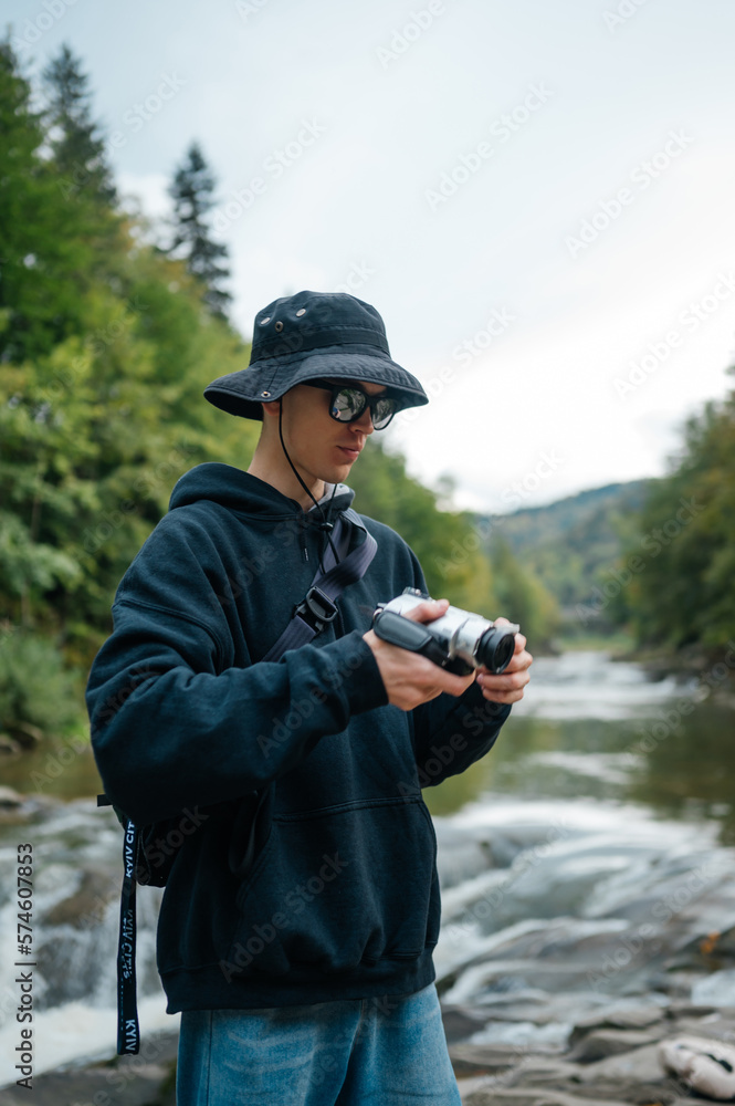 Stylish hipster man takes a photo on an old retro camera while hiking in the mountains against the background of a mountain river. Vertical