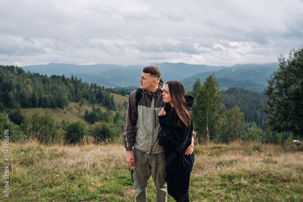 A beautiful couple of tourists are standing in the mountains during a hike against the background of beautiful views, hugging and looking away.