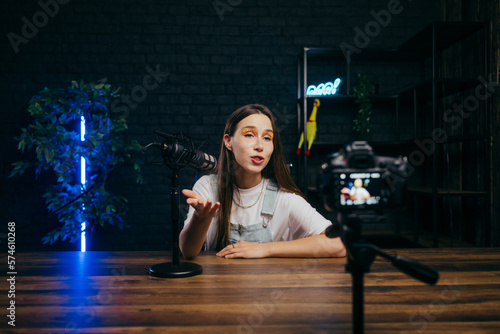 Backstage photo of female blogger sitting with microphone in cozy studio with blue light and recording video on camera, talking into studio microphone.