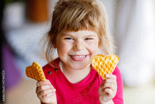 Portrait of happy little preschool girl holding fresh baked heart waffle. Smiling hungry toddler child with sweet biscuit wafer. Sweet sugar belgian waffles.