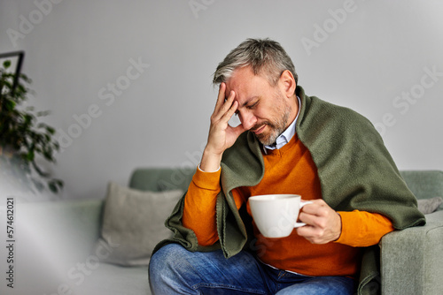 Obraz na płótnie A man holding his head, having a fever, drinking tea, and sitting on the couch