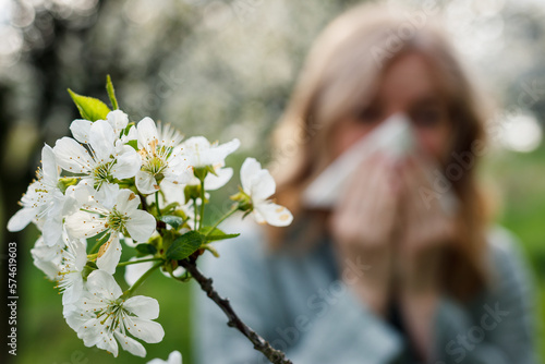 Woman sneezing and blowing nose in blooming park. Spring pollen allergy and hay fever. Selective focus on blossom photo