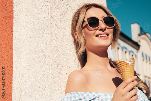 Young beautiful smiling hipster woman in trendy summer dress clothes. Sexy carefree model posing in the street. Positive blond female eating tasty ice cream in waffles cone in sunglasses at sunset