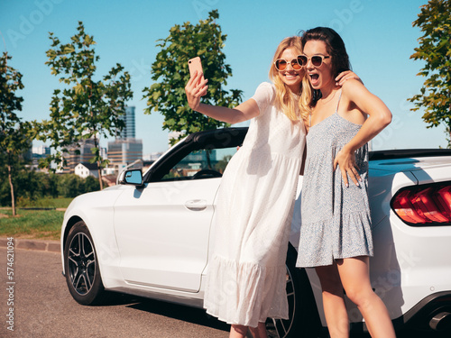 Two young beautiful and smiling hipster female in convertible car. Sexy carefree women posing near cabriolet. Positive models riding and having fun in sunglasses outdoors. Taking selfie photos © halayalex
