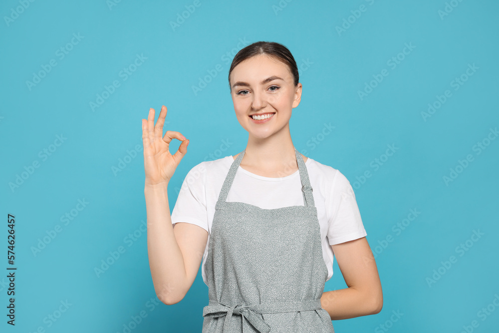 Beautiful young woman in clean apron with pattern on light blue background Beautiful young woman in clean apron with pattern on light blue background