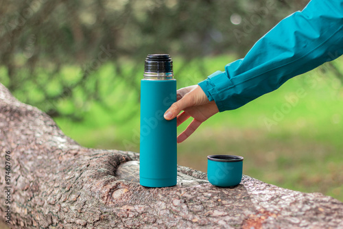 a human hand holds a steel green thermos, a mug with hot tea nearby on a branch of an old coniferous tree in the forest. Nature, picnic, camping