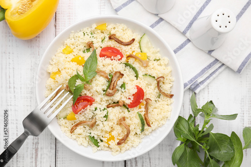 Bowl of delicious couscous with vegetables and basil served on white wooden table, flat lay