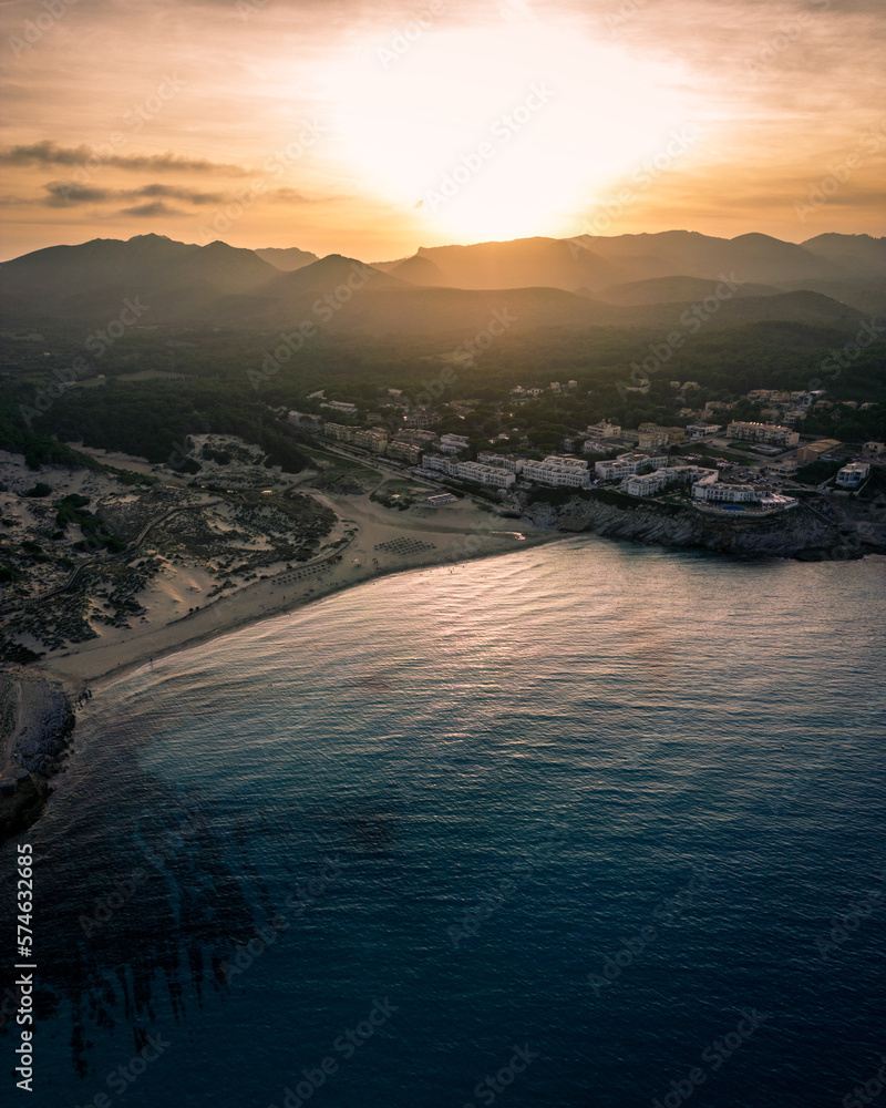 View over the shoreline of Mallorca, Spain, on a sunny afternoon in Summer showing a village by the sea with a beachduring blue hour facing the sun