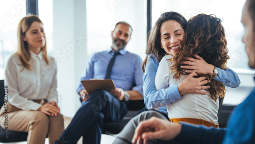 Support group gathering for a meeting. Two people are embracing each other and other members are supporting them. A multi-ethnic group of adults are attending a group therapy session.