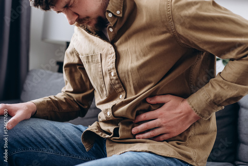 Upset male squeezing belly with hand because of abdominal pain. Unhappy young man sit on grey couch at home, suddenly feeling strong stomach ache, gastritis problem. Guy suffering from stomach ache photo