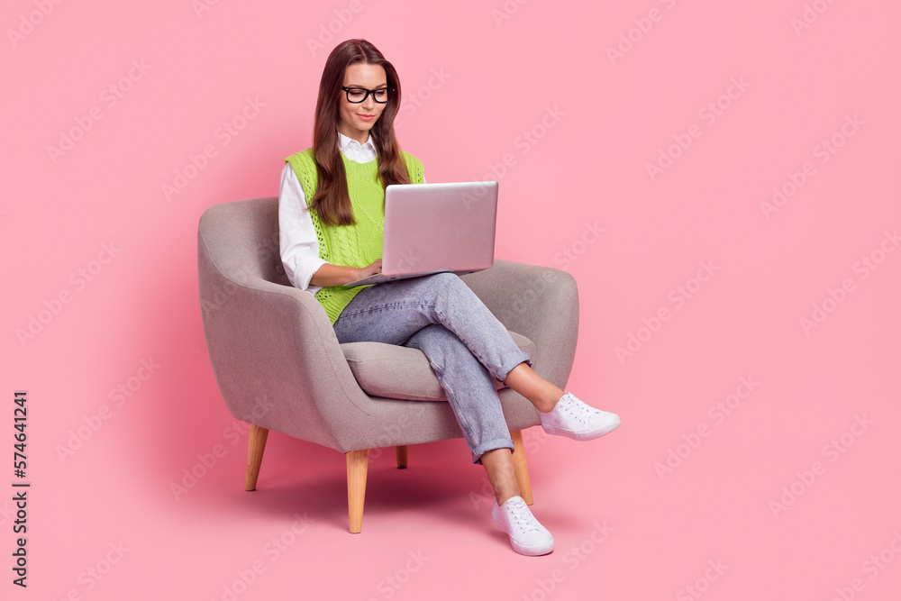 Full size photo of clever optimistic smart woman wear waistcoat jeans sit on armchair look at laptop isolated on pink color background Full size photo of clever optimistic smart woman wear waistcoat jeans sit on armchair look at laptop isolated on pink color background