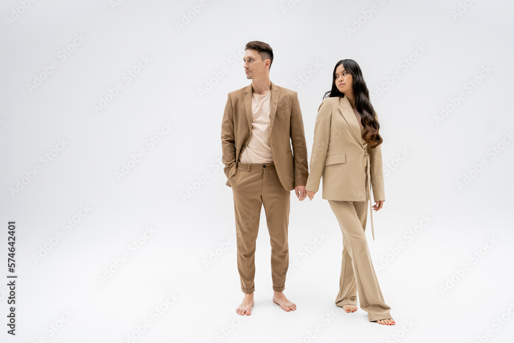 full length of barefoot interracial couple in beige pantsuits looking away on grey background. full length of barefoot interracial couple in beige pantsuits looking away on grey background.
