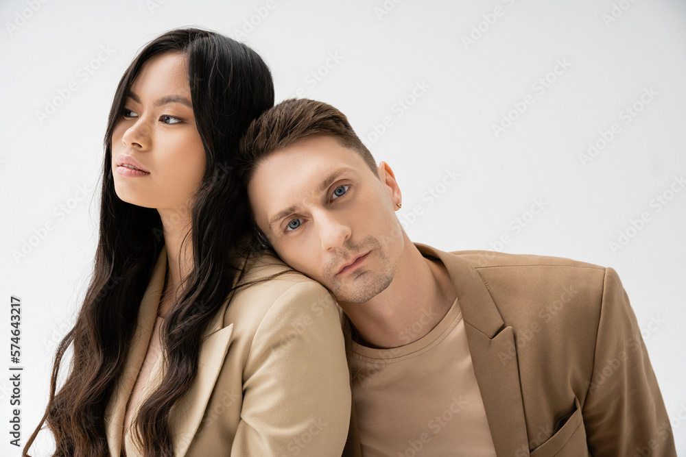 stylish man in beige blazer leaning on young asian woman with long brunette hair isolated on grey. stylish man in beige blazer leaning on young asian woman with long brunette hair isolated on grey.