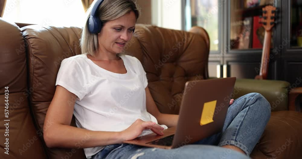 Female freelancer sits on sofa and uses laptop and headphones Female freelancer sits on sofa and uses laptop and headphones