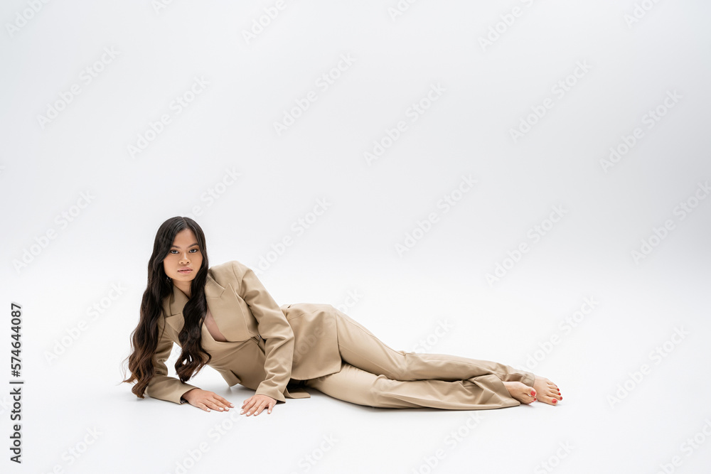 full length of barefoot asian model in fashionable suit lying and looking at camera on grey background. full length of barefoot asian model in fashionable suit lying and looking at camera on grey background.