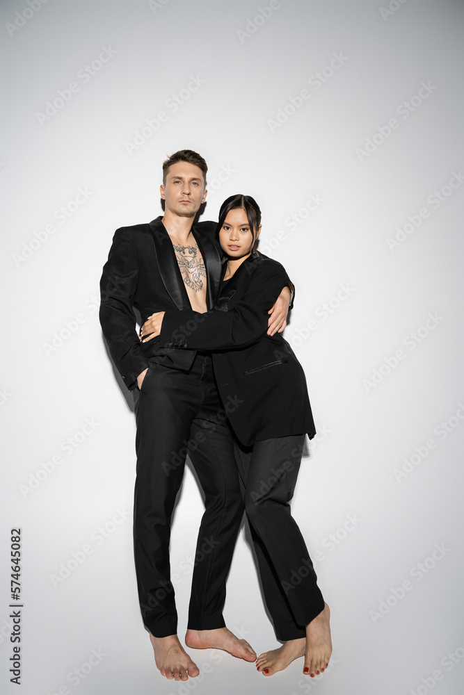full length of barefoot interracial couple in black elegant outfit embracing and looking at camera on grey background. full length of barefoot interracial couple in black elegant outfit embracing and looking at camera on grey background.