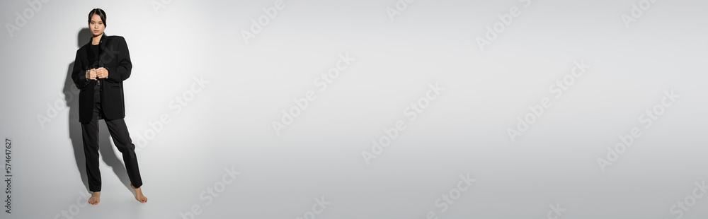 full length of barefoot asian woman in black pantsuit standing on grey background with shadow, banner. full length of barefoot asian woman in black pantsuit standing on grey background with shadow, banner.