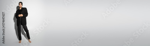 full length of barefoot asian woman in black pantsuit standing on grey background with shadow, banner.