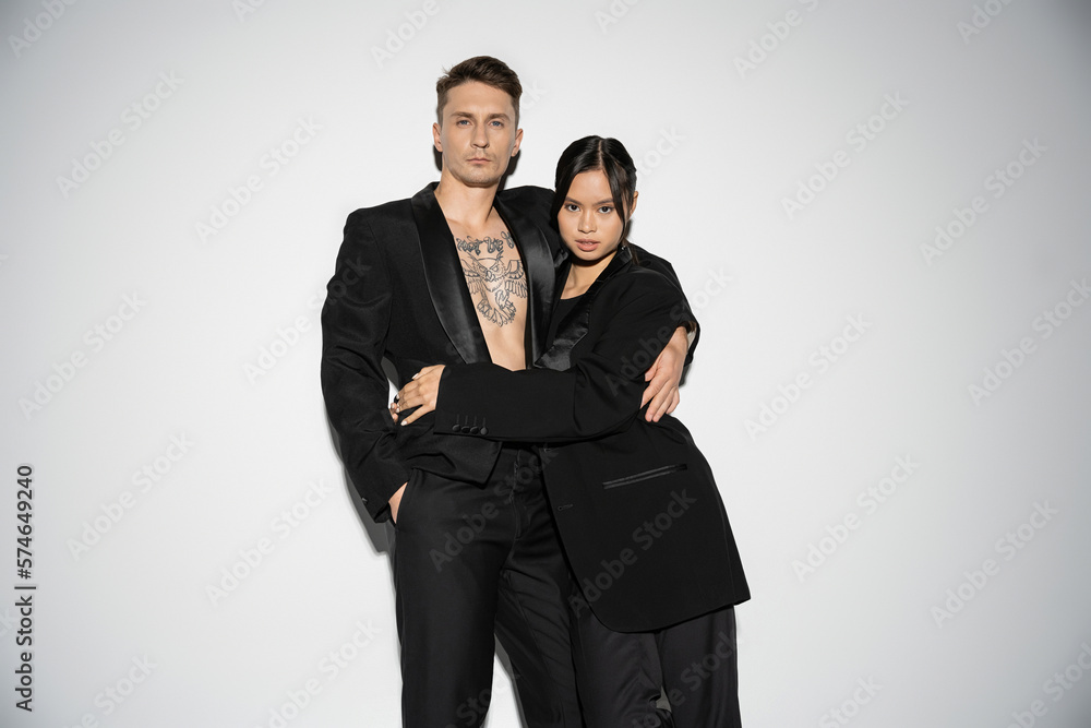 fashionable multiethnic couple in black elegant suits embracing and looking at camera on grey background. fashionable multiethnic couple in black elegant suits embracing and looking at camera on grey background.