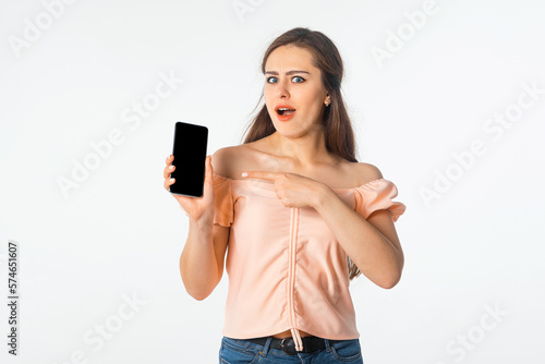 Surprised young woman in stylish outfit, pointing at mobile phone screen, showing smartphone display and looks amazed and impressed by smth, white background