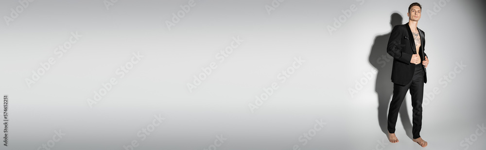 full length of barefoot tattooed man in black elegant pantsuit posing on grey background with shadow, banner. full length of barefoot tattooed man in black elegant pantsuit posing on grey background with shadow, banner.