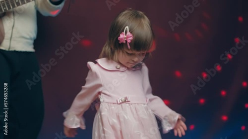 Little girl on stage in pink vintage dress, she dances, her father plays acoustic guitar. Color music is shining. Performance on stage. Early development of children