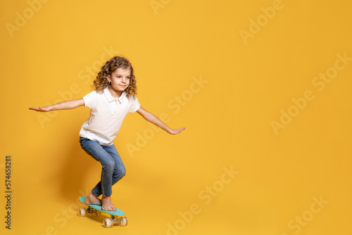 Fototapeta Naklejka Na Ścianę i Meble -  Full length of little girl in white polo, blue jeans who skating on penny board on yellow background