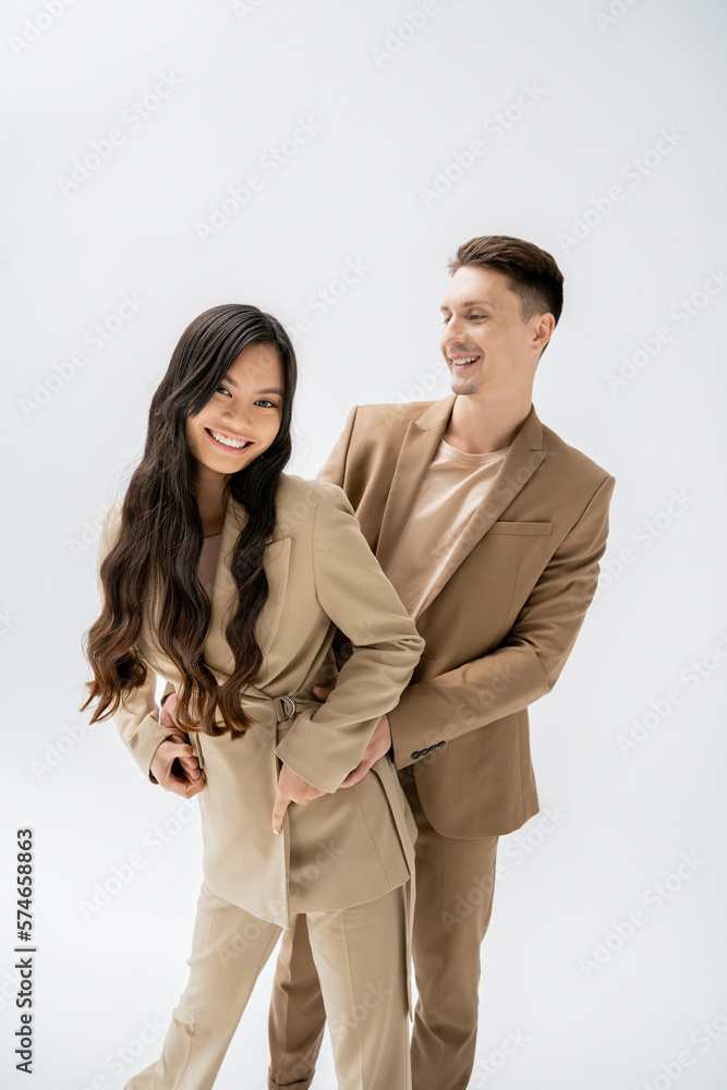 cheerful asian woman with long brunette hair looking at camera near trendy man isolated on grey. cheerful asian woman with long brunette hair looking at camera near trendy man isolated on grey.