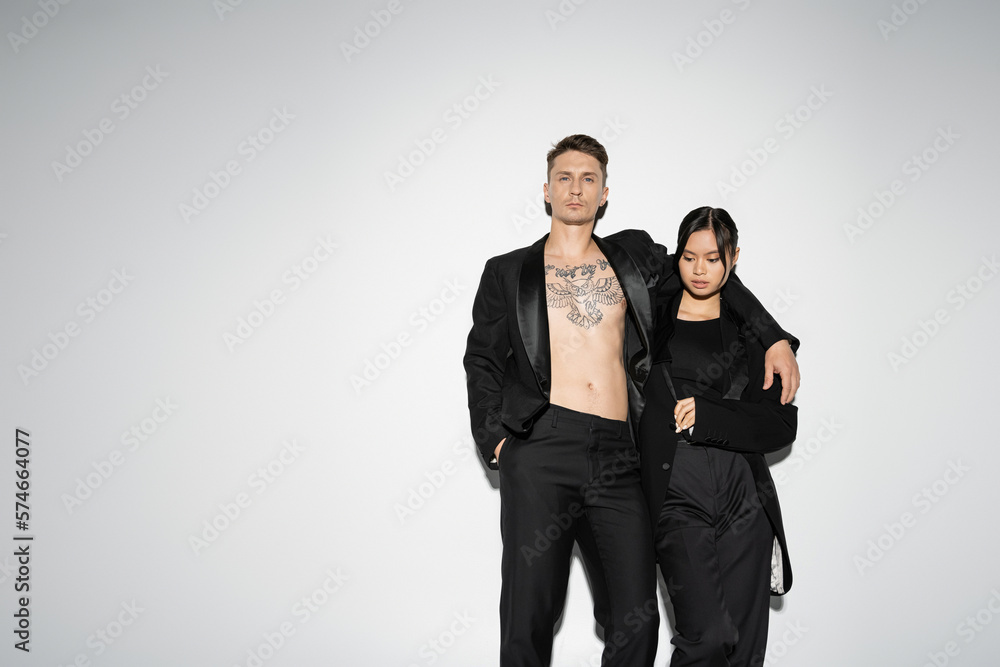 tattooed man in black trendy suit embracing elegant asian woman while posing with hand in pocket on grey background.