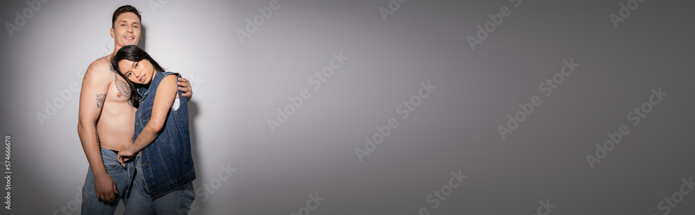 shirtless man with tattooed body embracing brunette asian woman in denim vest on grey background, banner.