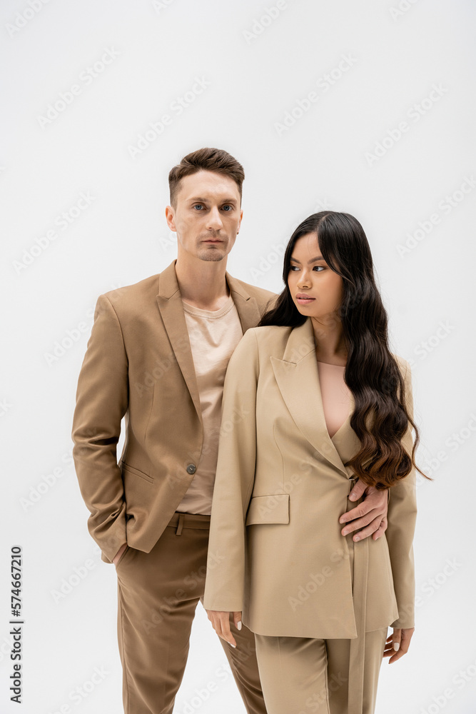 man in beige suit standing with hand in pocket and embracing asian woman isolated on grey. man in beige suit standing with hand in pocket and embracing asian woman isolated on grey.