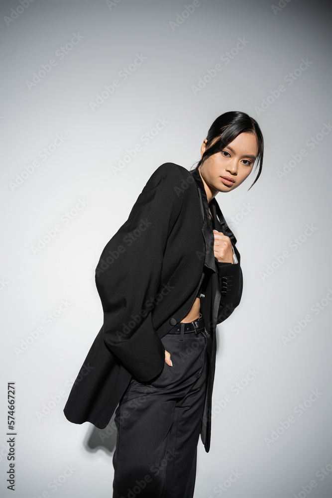 Fototapeta premium brunette asian woman in black elegant blazer and trousers standing with hand in pocket on grey background.