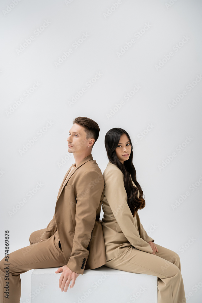 brunette asian woman with long hair looking at camera while sitting back to back with trendy man isolated on grey. brunette asian woman with long hair looking at camera while sitting back to back with trendy man isolated on grey.