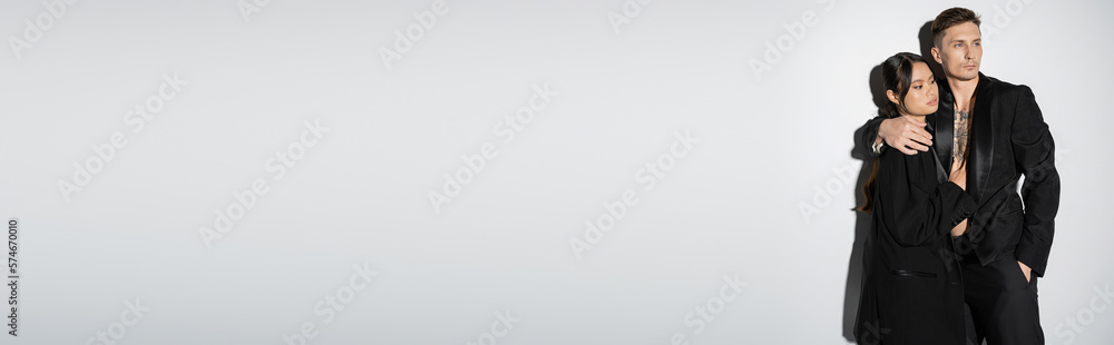 interracial couple in black fashionable outfit looking away on grey background, banner. interracial couple in black fashionable outfit looking away on grey background, banner.