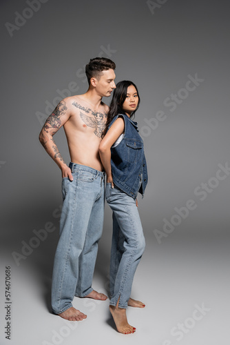 full length of barefoot tattooed man in jeans near brunette asian woman in denim outfit posing on grey background.