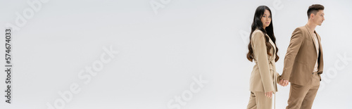 фотографія brunette asian woman looking at camera and holding hands with man in trendy suit isolated on grey, banner