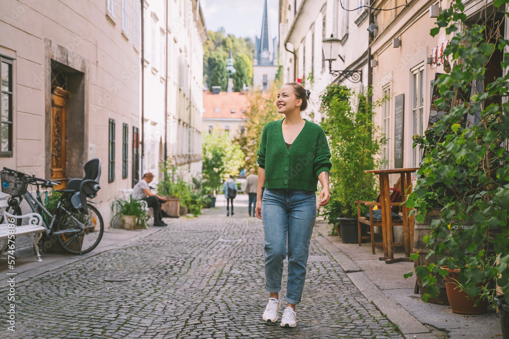 Obraz na płótnie Photo of friendly smiling attractive woman on background of green hedge in the street of european city. Happy confident woman in front of green leaves near buildings. w salonie