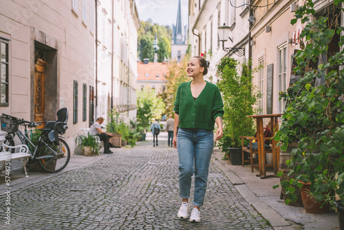 Photo of friendly smiling attractive woman on background of green hedge in the street of european city. Happy confident woman in front of green leaves near buildings.