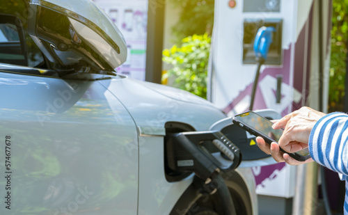 Woman using smartphone paying service in app on blur EV car charging at electric car charging station. EV car charging point. Electric vehicle fast charge. Sustainable power for reduce carbon emission