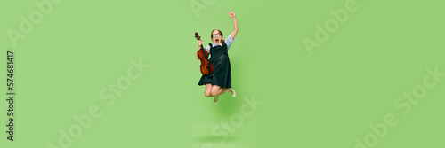 Cute little girl, talanted musician wearing huge mother's heels shoes jumping with violin, having fun isolated over green background. Banner with copy space for ad photo