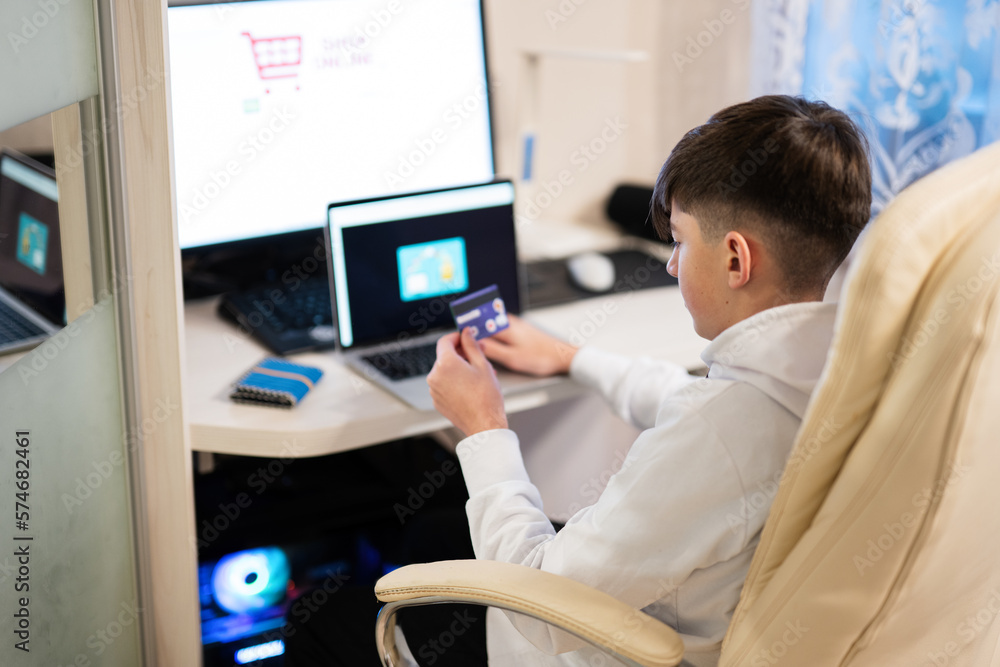 Fototapeta premium Happy teenage boy using laptop and credit card, sitting at desk and ordering goods online from home.