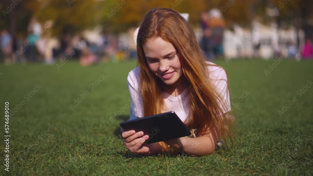 Teen girl on green lawn in park lying with a tablet in her hands doing her studies. Realtime