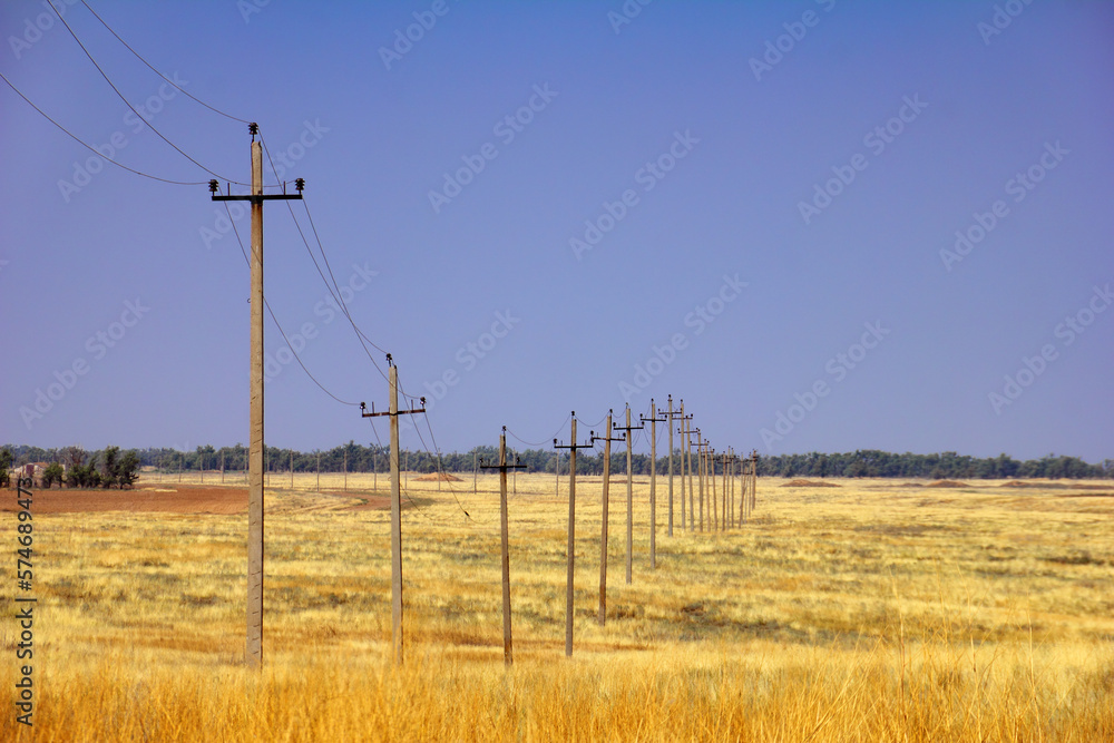 A row of power line supports on the steppe field with dry yellow grass at the background of blue evening sky and distant wood. Beautiful scenic view in Kalmykia, surroundings of Elista, South Russia