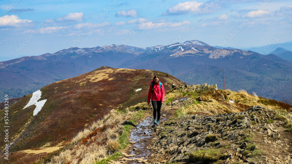 girl in pink jacket walks along a mountain ridge during spring; mountain walk with vegetation coming to life, colourful spring in the mountains