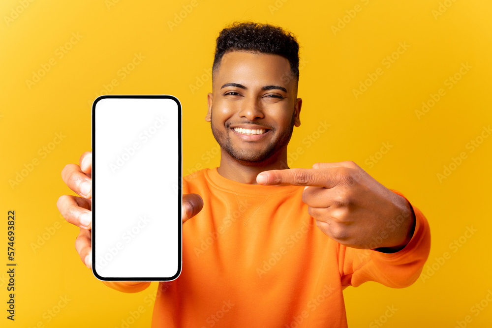 Surprised Indian man holding smartphone in hand and points finger at empty phone screen, astonishment guy presenting best deal, advertising new mobile app, isolated on yellow - obrazy, fototapety, plakaty 