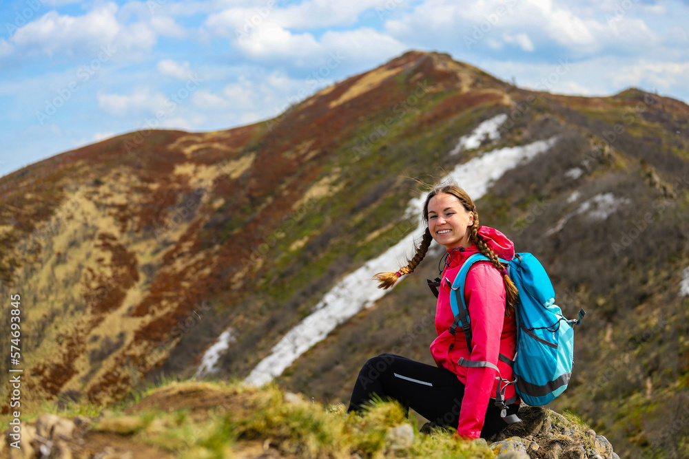 beautiful hiker girl with backpack sitting on the grass with large colorful mountain in front of her; hiking in the mountains during early spring