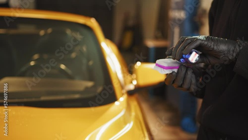 Cropped image of hands of male auto service worker in protective gloves, applying special polish cream, nano-ceramic coating on sponge, to polish yellow luxury car on the background. Car detailing photo